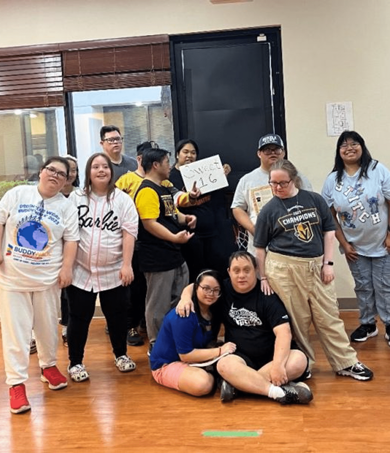 A diverse group of smiling people pose in a room, one holding a sign that reads "Sweet 16".